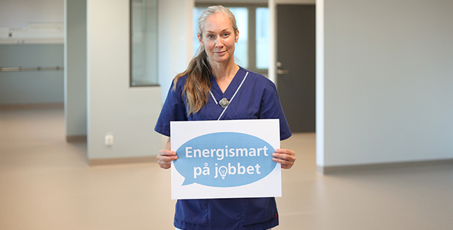 Kvinna i vårdkläder som håller i en skylt där det står Energismart på jobbet. 